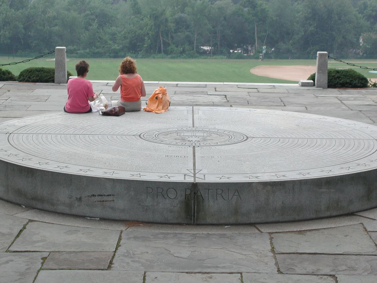 Two women sit, presumably eating together, on a short, round, wide sculpture in stone showing markings of a compass.